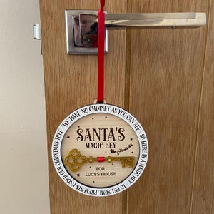 May include: A white round wooden ornament with the text "We have no chimney as you can see, so here is a magic key. Santa's Magic Key for Lucy's House. Put some presents under our Christmas tree." and a gold glitter key.