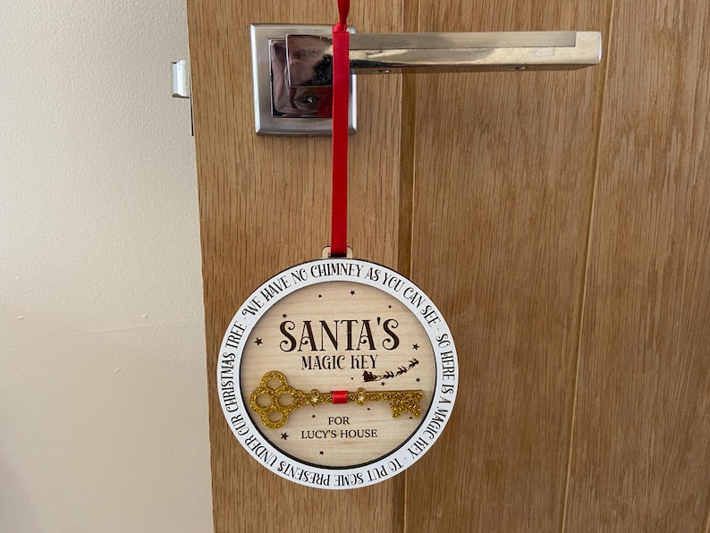 May include: A white round wooden ornament with the text "We have no chimney as you can see, so here is a magic key. Santa's Magic Key for Lucy's House. Put some presents under our Christmas tree." and a gold glitter key.