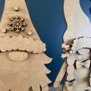 May include: Two wooden gnome ornaments, one with a snowflake on its hat and the other with a braid and bows. The gnomes are unfinished and ready to be painted.