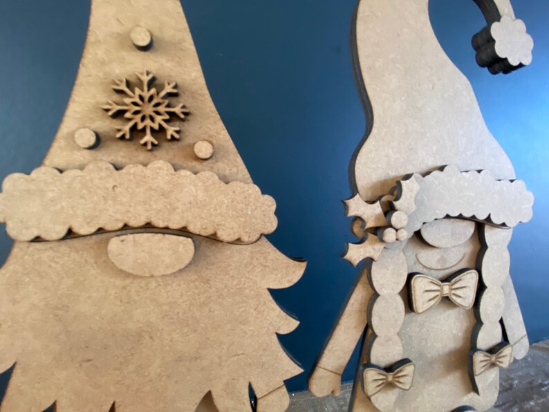May include: Two wooden gnome ornaments, one with a snowflake on its hat and the other with a braid and bows. The gnomes are unfinished and ready to be painted.