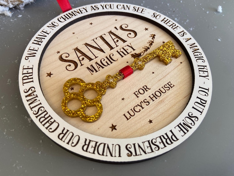 May include: A round wooden ornament with a gold glitter key and the text "Santa's Magic Key" and "For Lucy's House". The ornament also has the text "We have no chimney as you can see - so here is a magic key - to put some presents under our Christmas tree".