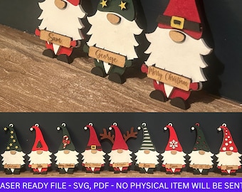 Puede incluir: Un conjunto de 10 adornos de gnomo de madera cortados con láser con varios diseños navideños. Los gnomos están pintados en rojo, verde y blanco con detalles negros. Cada gnomo tiene un diseño diferente en su gorro, incluyendo estrellas, bastones de caramelo, copos de nieve y cuernos de reno. Los gnomos sostienen carteles con el texto "Feliz Navidad", "Sam", "John", "George", y "Santa".