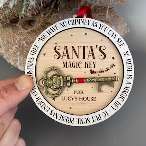 May include: A round wooden ornament with a white border and the text "We have no chimney as you can see - so here is a magic key to put some presents under our Christmas tree. Santa's Magic Key for Lucy's House". A gold-colored key with a red ribbon is attached to the ornament.