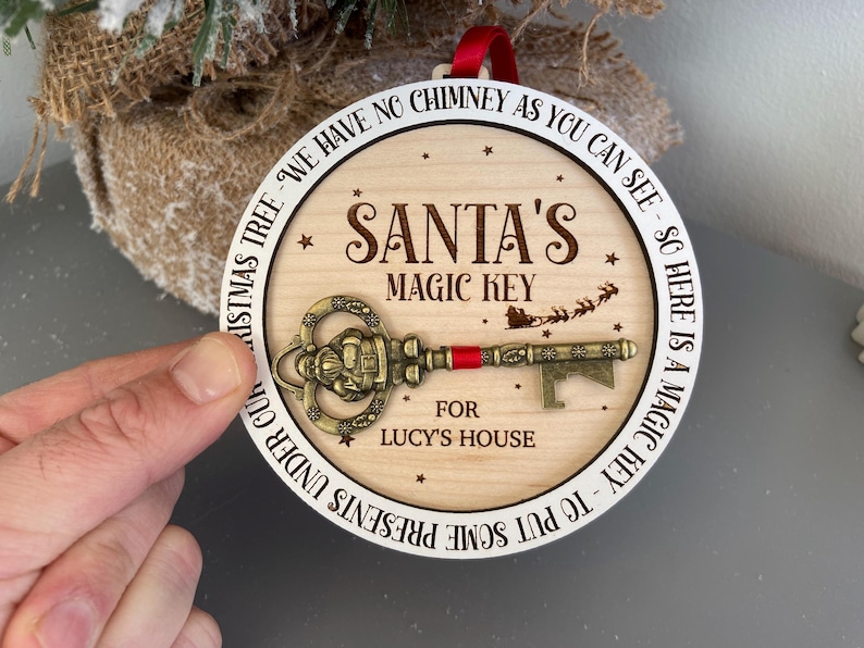 May include: A round wooden ornament with a white border and the text "We have no chimney as you can see - so here is a magic key to put some presents under our Christmas tree. Santa's Magic Key for Lucy's House". A gold-colored key with a red ribbon is attached to the ornament.