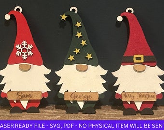 Puede incluir: Tres adornos de gnomo de Navidad de madera pintados en rojo, verde y blanco. Los gnomos tienen diferentes diseños en sus sombreros, incluyendo un copo de nieve, estrellas y un gorro de Papá Noel. Los adornos tienen nombres personalizados: Sam, George y Feliz Navidad.