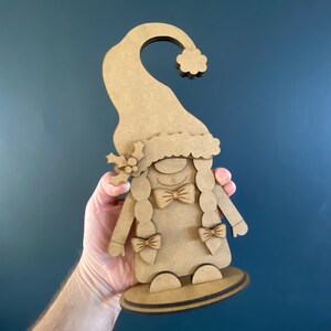 May include: A wooden cutout of a Christmas gnome with a red hat, white beard, and a bow tie. The gnome is holding a small sprig of holly in its hand.