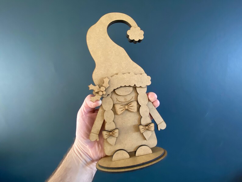 May include: A wooden cutout of a Christmas gnome with a red hat, white beard, and a bow tie. The gnome is holding a small sprig of holly in its hand.