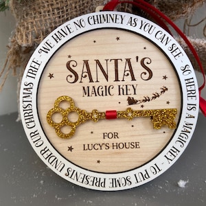 May include: A round wooden ornament with a white border and gold glitter key. The ornament reads "We have no chimney as you can see - so here is a magic key - to put some presents under our Christmas tree - Santa's Magic Key for Lucy's House".