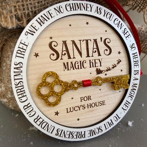 May include: A round wooden ornament with a white border and the text "We have no chimney as you can see - so here is a magic key to put some presents under our Christmas tree - Santa's Magic Key for Lucy's House". A gold glitter key is attached to the ornament.