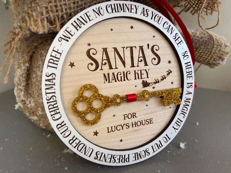 May include: A round wooden ornament with a white border and the text "We have no chimney as you can see - so here is a magic key to put some presents under our Christmas tree - Santa's Magic Key for Lucy's House". A gold glitter key is attached to the ornament.