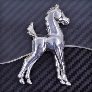 Puede incluir: Figura de caballo de metal plateado. El caballo está representado de pie con las patas ligeramente separadas. La figura tiene una melena y una cola detalladas. El fondo es una superficie oscura y texturizada.
