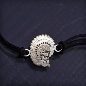 Pode incluir: Pulseira prateada com um pendente detalhado de cocar nativo americano. O pendente está ligado a uma pulseira de cordão preto com extremidades ajustáveis. O design apresenta detalhes intrincados.
