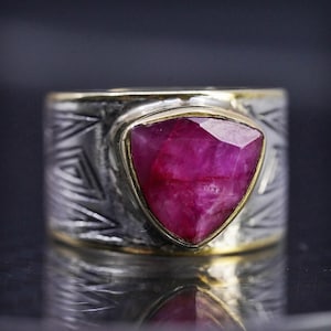 May include: A wide band ring featuring a triangular, faceted deep pink gemstone set in a gold-toned bezel. The silver-toned band has a textured, oxidized finish with engraved geometric triangular patterns wrapping around the entire circumference.