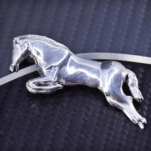 Puede incluir: Una figurita de caballo de color plateado, saltando hacia adelante con las patas extendidas. La figurita está detallada con una melena y una cola esculpidas, y se coloca sobre un fondo negro.