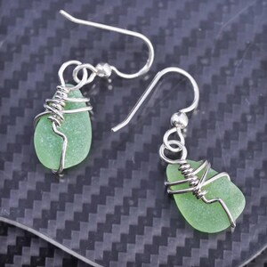 May include: A pair of handcrafted earrings featuring sea glass in a soft green hue. Each piece of sea glass is wrapped with silver-colored wire and suspended from a silver-colored hook.