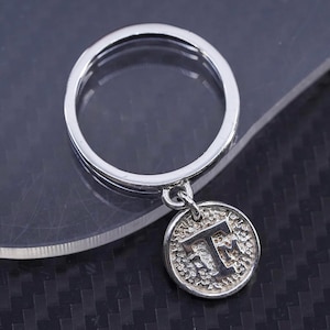 May include: A silver ring with a circular band and a small, round pendant. The pendant features the letter "T" in a textured design. The ring is displayed on a dark surface.
