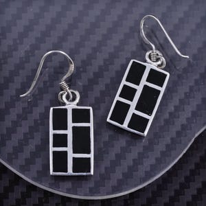 May include: A pair of silver dangle earrings with black rectangular pendants. The pendants have a geometric design of squares.