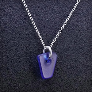 May include: A silver chain necklace with a pendant made of blue sea glass. The pendant is a small, irregular shape with a hole near the top. The necklace is displayed against a dark background.