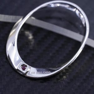 Puede incluir: Colgante ovalado plateado pulido con una pequeña gema roja redonda engastada cerca de la base. El colgante está marcado "925 ELLE CHINA". El diseño es liso y moderno, reflejando la luz.