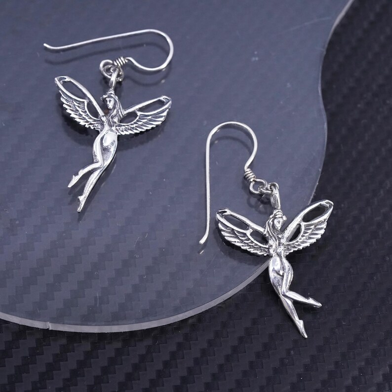 K&ouml;nnte beinhalten: Ein Paar silberne Ohrringe mit einem gefl&uuml;gelten Figuren-Design. Die Ohrringe sind im H&auml;nge-Stil mit einem Hakenverschluss.