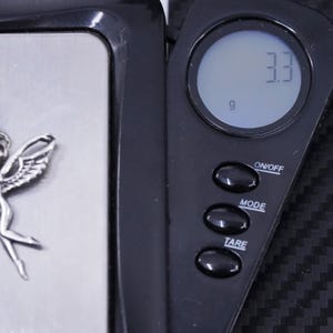 K&ouml;nnte beinhalten: Eine schwarze digitale Waage mit einer silbernen Wiegeplattform. Die Waage zeigt "3,3 g" auf dem Bildschirm an. Die Waage hat Tasten mit den Beschriftungen "ON/OFF", "MODE" und "TARE".