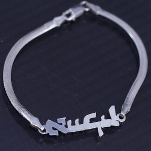 May include: A silver-toned herringbone chain bracelet featuring a central nameplate with stylized, angular lettering. The bracelet includes a secure lobster claw clasp mechanism, creating a continuous, polished metallic loop design.