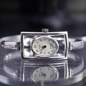 May include: Silver-toned quartz watch with an oval face and a rectangular frame. The watch face is white with black numerals and hands. The watch band is a silver bangle style.