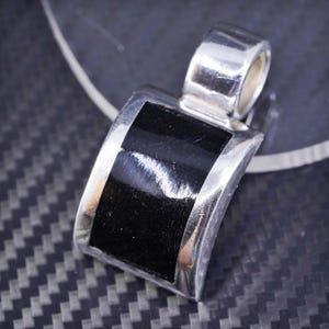 May include: A silver pendant with a black rectangular inlay. The pendant has a curved shape and a loop at the top for attaching a chain.