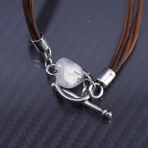 May include: A close-up of a brown leather bracelet with a silver clasp. The bracelet features multiple strands of brown leather and a clear, heart-shaped stone. The clasp is a toggle style, with a silver bar and loop.