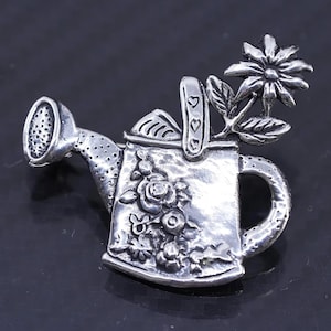 Puede incluir: Un broche de plata en forma de regadera con un diseño floral. La regadera tiene un asa y un pico. La flor tiene un diseño detallado con pétalos y hojas. El broche es un gran regalo para los jardineros o cualquier persona que ame las flores.