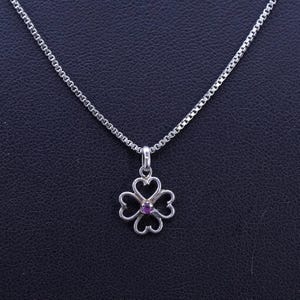 May include: A silver necklace with a clover pendant. The pendant features four heart-shaped leaves surrounding a small, round, purple gemstone. The chain is a delicate box chain style, and the background is a dark, textured surface.