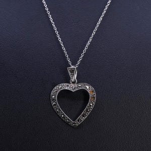 Puede incluir: Collar con colgante de corazón de plata. El colgante tiene forma de corazón con el centro hueco y está incrustado con pequeñas piedras oscuras. La cadena del collar es delicada y plateada. El fondo es una superficie oscura y texturizada.