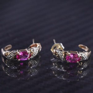 May include: A pair of gold-toned hoop earrings. Each earring features a central oval-shaped pink gemstone, surrounded by small, clear accent stones. The earrings have a curved design with decorative detailing.