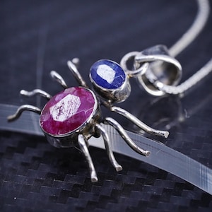 May include: A silver spider-shaped pendant necklace featuring a large, oval-cut, faceted deep pink ruby-colored stone for the body and a smaller, oval-cut, faceted deep blue sapphire-colored stone for the head, with eight thin silver legs.