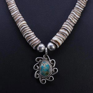 Puede incluir: Un collar artesanal con un colgante turquesa y filigrana plateada. El colgante cuelga de una hebra de cuentas de concha blancas y marrones, conectadas por cuentas plateadas. El collar se exhibe sobre una superficie negra.