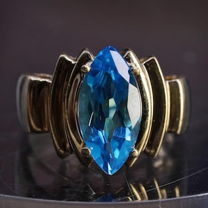 Puede incluir: Un anillo de tono dorado con una gran piedra azul de talla marquesa facetada en el centro. La banda tiene un diseño ancho y estructurado con crestas verticales a ambos lados de la piedra central, creando una apariencia geométrica audaz.