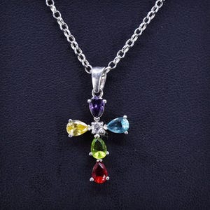 May include: A silver chain necklace with a cross pendant featuring a rainbow of gemstones. The cross is adorned with a clear stone in the center and pear-shaped stones in various colors, including red, orange, yellow, green, blue, and purple.
