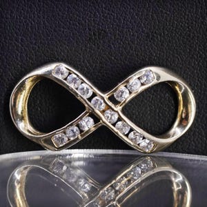 May include: A gold-toned infinity symbol pendant, encrusted with small, clear gemstones. The pendant is displayed on a reflective surface against a dark background, highlighting its shape and sparkle. The design symbolizes eternity.