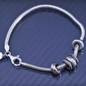 Puede incluir: Un brazalete de cadena de plata con un dije de corazón y tres cuentas de plata. Las cuentas están grabadas con la palabra "Friends".