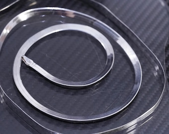 Puede incluir: Un collar de cadena de espiga plateada enrollado sobre una superficie oscura. El collar tiene un diseño plano y tejido y un brillo sutil. El cierre es visible al final de la cadena. La joya se exhibe sobre un fondo texturizado.