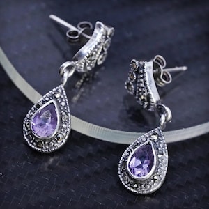 May include: A pair of silver drop earrings with a pear-shaped, light purple gemstone center. The earrings have a marcasite border and a post-back closure. The earrings are displayed on a dark surface.