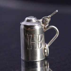 May include: Silver charm in the shape of a beer stein with a hinged lid and a handle. The stein has the initials "HB" engraved on the front.