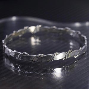 Peut inclure: Un bracelet argenté au design torsadé. Le bracelet est composé de maillons rectangulaires reliés par des accents métalliques torsadés. Le bijou est présenté sur une surface réfléchissante.