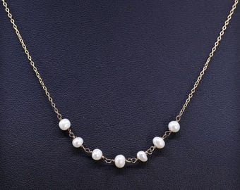 Puede incluir: Un delicado collar de cadena dorada con una serie de perlas blancas. Las perlas están espaciadas uniformemente a lo largo de la cadena, creando un diseño sutil y elegante. El collar se muestra sobre un fondo oscuro.