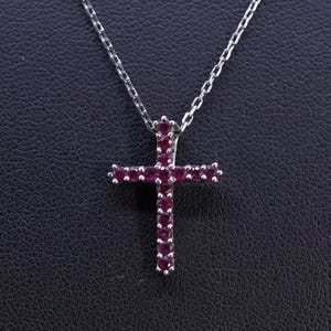 Puede incluir: Un collar plateado con un colgante de cruz. La cruz está adornada con numerosas piedras preciosas pequeñas de color rojo intenso. La cadena es delicada y el colgante está centrado sobre un fondo oscuro, resaltando el diseño de la joya.