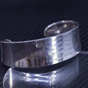 May include: A silver-toned cuff bracelet with engraved script. The text reads "May you walk in Peace, Live with Love, Work with Joy, and May Your God go with A Simple Blessing."
