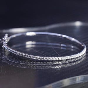 May include: A silver bangle bracelet with a row of small, clear gemstones set along the top. The bracelet is round and has a clasp. The background is dark, and the bracelet is reflected in the surface.
