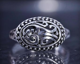 May include: Silver ring with an oval, openwork design. The ring has an intricate, swirling pattern within the oval and a beaded border. The ring is shown on a reflective surface.