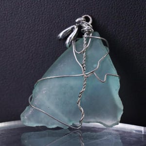 May include: A sea glass pendant with a light blue triangular shape. The sea glass is wrapped in silver-colored wire, with a twisted wire bail at the top. The pendant is displayed against a dark background.