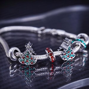 Puede incluir: Un brazalete de plata con tres dijes de árbol de Navidad. Los dijes están decorados con cristales rojos y verdes.